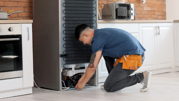 Professional technician using tools to fix refrigerator compressor and coils in a kitchen.