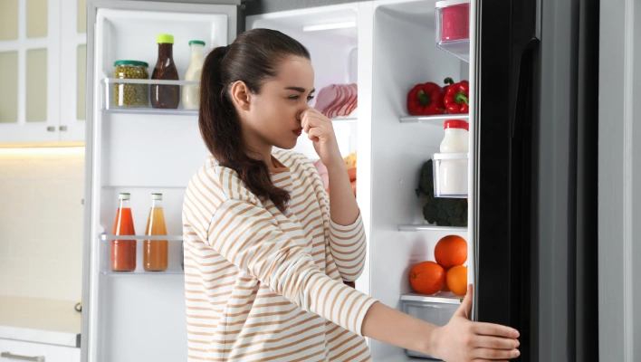 A disappointed woman pinching her nose while opening a clean kitchen appliance because the fridge smells bad even after maintenance.