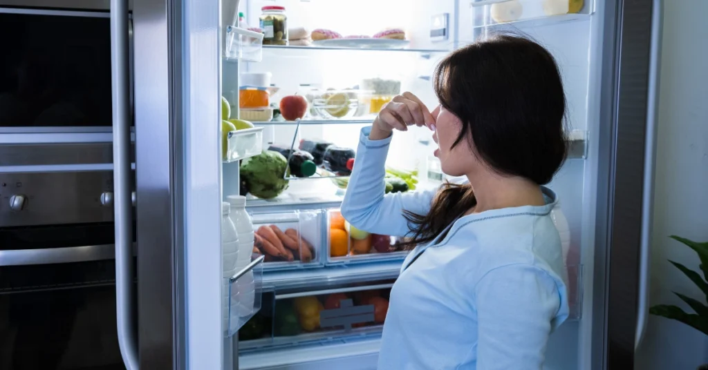A woman holding her nose while looking inside an open side-by-side refrigerator because the fridge smell bad.